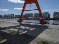 an orange crane is sitting in the field by some water and buildings on a sunny day