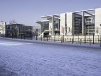 snow covering the ground and sidewalk of an industrial area with buildings, fences, and benches