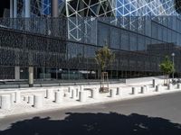 concrete street posts along the street side are lined with plants and trees in front of a glass building