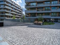 the empty bench is near an outdoor courtyard area of a large building with high rise apartment buildings