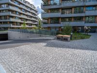 the empty bench is near an outdoor courtyard area of a large building with high rise apartment buildings