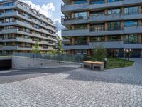 the empty bench is near an outdoor courtyard area of a large building with high rise apartment buildings