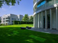 the lawn is green and features a stone bench, grass, large buildings, and a tree