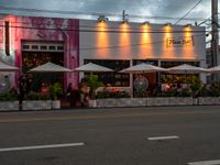 the front of an establishment with tables and umbrellas sitting outside it on a street