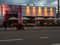 the front of an establishment with tables and umbrellas sitting outside it on a street