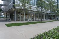 the concrete walkway has grass in front of it and several tables are outside by the building