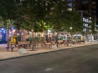 a city street lined with tables and chairs under a string light at night with people walking on the sidewalk