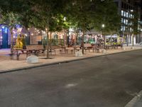 a city street lined with tables and chairs under a string light at night with people walking on the sidewalk