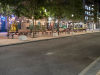 a city street lined with tables and chairs under a string light at night with people walking on the sidewalk