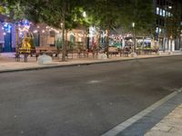 a city street lined with tables and chairs under a string light at night with people walking on the sidewalk