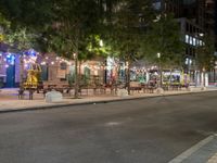 a city street lined with tables and chairs under a string light at night with people walking on the sidewalk