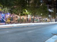 a city street lined with tables and chairs under a string light at night with people walking on the sidewalk