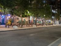 a city street lined with tables and chairs under a string light at night with people walking on the sidewalk