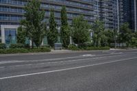 a tall building next to an empty road with trees in front of it, and a row of parked cars beside