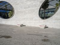 two white benches sit at the base of a large building on concrete and two circular windows sit at each end