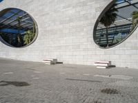 two white benches sit at the base of a large building on concrete and two circular windows sit at each end