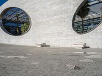 two white benches sit at the base of a large building on concrete and two circular windows sit at each end