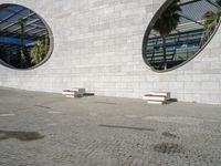 two small windows in an exterior with circular windows, a sidewalk and palm trees near by