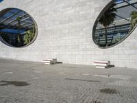 two small windows in an exterior with circular windows, a sidewalk and palm trees near by