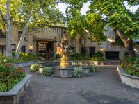 an outdoor fountain surrounded by flower gardens near two buildings and trees in the distance that includes plants and tree branches