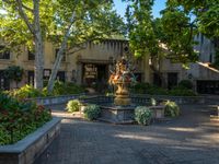 an outdoor fountain surrounded by flower gardens near two buildings and trees in the distance that includes plants and tree branches