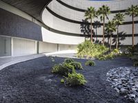 a courtyard is seen under a curved roof above shrubs and trees, the black pebbles look like volcanic