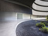 a courtyard is seen under a curved roof above shrubs and trees, the black pebbles look like volcanic