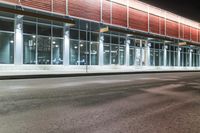 this is a bus stop at night next to the curb of a building with a large mirror window