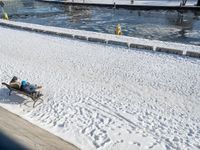 a person sitting on a bench in the snow next to a river and boats docked