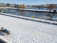 a person sitting on a bench in the snow next to a river and boats docked