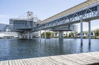 an empty pier sitting in front of large metal structures, with buildings along the bank of the river