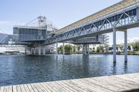 an empty pier sitting in front of large metal structures, with buildings along the bank of the river