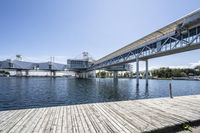 an empty pier sitting in front of large metal structures, with buildings along the bank of the river