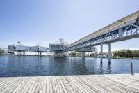 an empty pier sitting in front of large metal structures, with buildings along the bank of the river