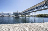 an empty pier sitting in front of large metal structures, with buildings along the bank of the river