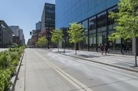 a paved sidewalk in front of tall buildings and trees in a city park, a couple people walk down the sidewalk to the left
