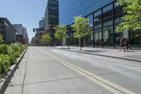 a paved sidewalk in front of tall buildings and trees in a city park, a couple people walk down the sidewalk to the left