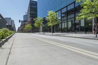 a paved sidewalk in front of tall buildings and trees in a city park, a couple people walk down the sidewalk to the left