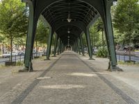 a walkway is lined with green metal supports and trees as cars pass in the distance