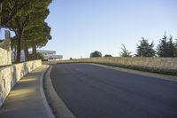 the roadway that runs along a stone wall by a sidewalk with traffic on it and two trees