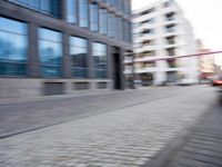 blurry photograph of a busy street with buildings and cars on the road and on brick path