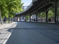 trees line the street in front of buildings with lots of windows and many vehicles parked under the bridge