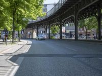 trees line the street in front of buildings with lots of windows and many vehicles parked under the bridge