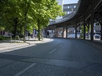 trees line the street in front of buildings with lots of windows and many vehicles parked under the bridge