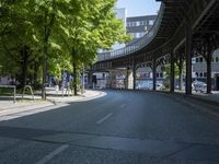 trees line the street in front of buildings with lots of windows and many vehicles parked under the bridge