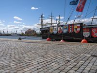 a row of flags and poles on the side of a dock with two ships in the back ground