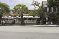 a cafe with lots of tables and umbrellas on a street corner near trees and a white fence