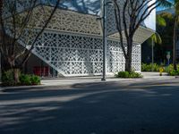 there is an empty street in the foreground, there is an open area for trees, bushes, and a white facade with small, latticed patterns on the walls