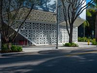 there is an empty street in the foreground, there is an open area for trees, bushes, and a white facade with small, latticed patterns on the walls