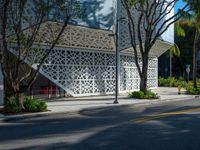 there is an empty street in the foreground, there is an open area for trees, bushes, and a white facade with small, latticed patterns on the walls